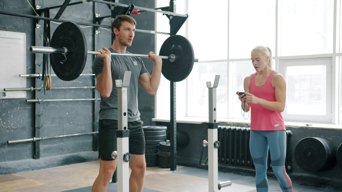 Man gör knäböj med skivstång i ett ljust gym medan en tränare tittar på